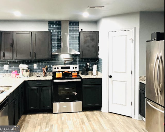 kitchen featuring appliances with stainless steel finishes, wall chimney range hood, decorative backsplash, light wood-style floors, and light stone countertops