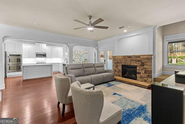 living room with a stone fireplace, hardwood / wood-style flooring, ceiling fan, arched walkways, and visible vents