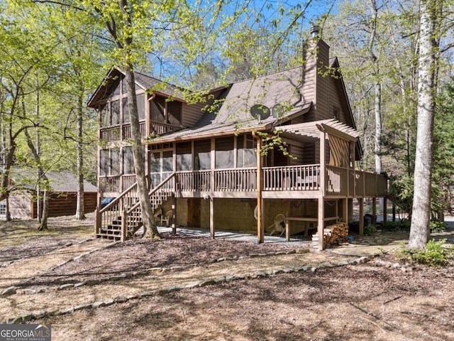 back of house featuring a chimney, stairs, a shingled roof, a sunroom, and a deck