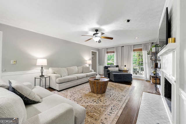 living area with wood-type flooring, a ceiling fan, a fireplace with flush hearth, and ornamental molding