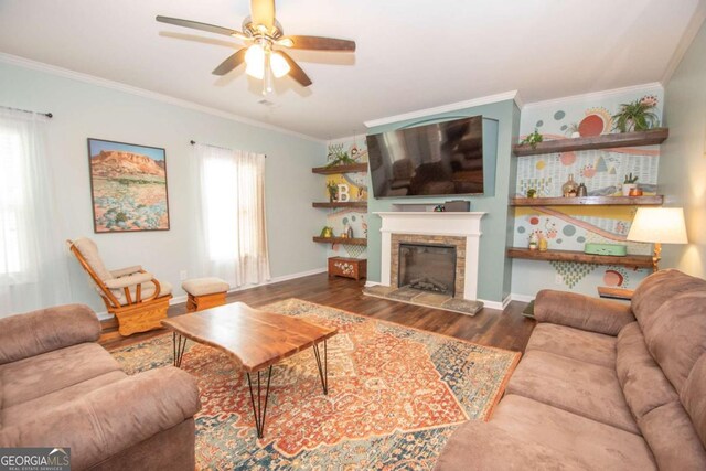 living area with crown molding, a stone fireplace, wood finished floors, ceiling fan, and baseboards