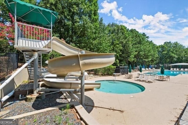 community pool with a patio area, a water slide, fence, and view of scattered trees