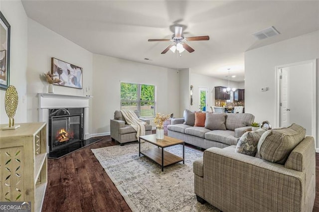 living area with visible vents, a glass covered fireplace, dark wood-style floors, and ceiling fan with notable chandelier