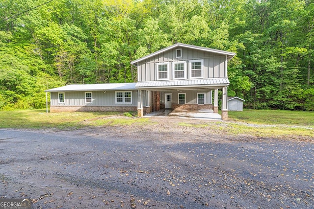 view of front of house with board and batten siding, a view of trees, covered porch, a storage unit, and driveway