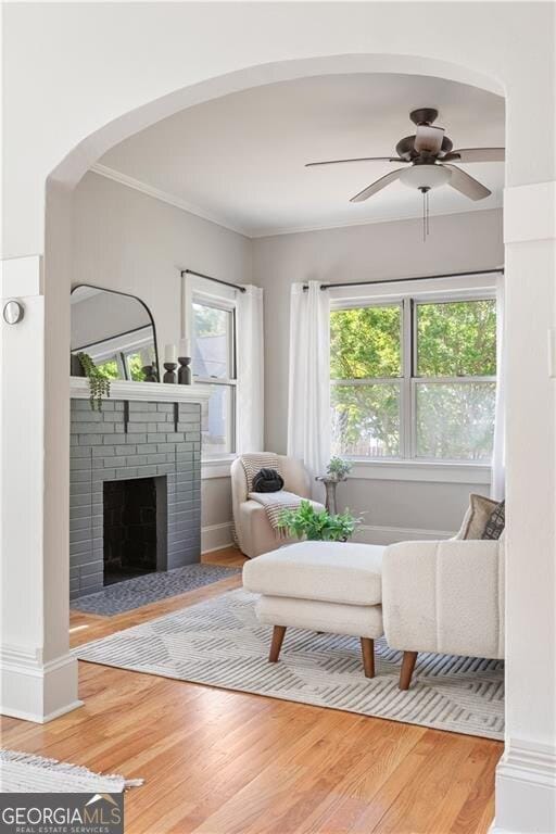 sitting room featuring crown molding, a ceiling fan, wood finished floors, arched walkways, and a brick fireplace