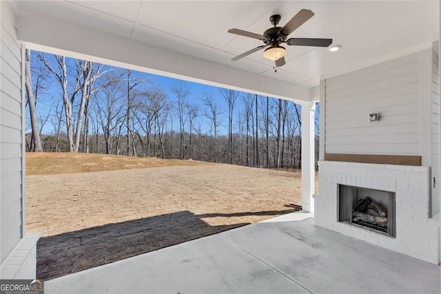 view of patio featuring ceiling fan, a view of trees, and an outdoor brick fireplace