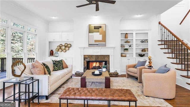 living room with a ceiling fan, wood finished floors, crown molding, a fireplace, and stairs