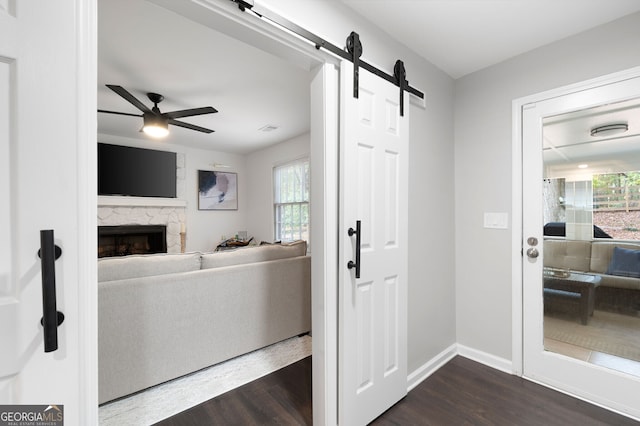 interior space featuring a stone fireplace, dark wood-type flooring, ceiling fan, and a barn door