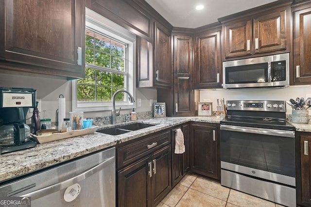 kitchen featuring stainless steel appliances, a sink, light tile patterned flooring, dark brown cabinetry, and light stone counters