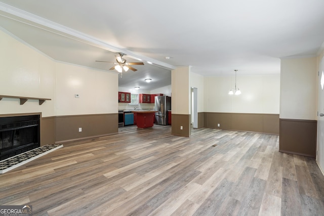unfurnished living room with a fireplace with flush hearth, a wainscoted wall, light wood-type flooring, ceiling fan with notable chandelier, and crown molding