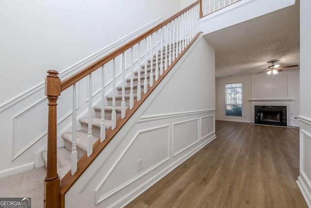staircase featuring a wainscoted wall, a ceiling fan, wood finished floors, a decorative wall, and a fireplace
