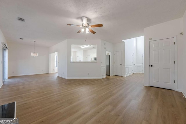 unfurnished living room with a textured ceiling, light wood-type flooring, baseboards, visible vents, and ceiling fan with notable chandelier