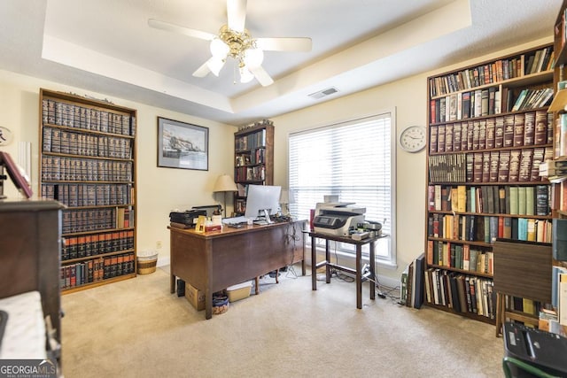 carpeted office with ceiling fan, baseboards, a tray ceiling, and visible vents