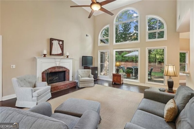 living area with a ceiling fan, a towering ceiling, a brick fireplace, healthy amount of natural light, and wood finished floors