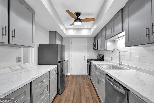 kitchen featuring gray cabinets, a ceiling fan, appliances with stainless steel finishes, a sink, and a tray ceiling