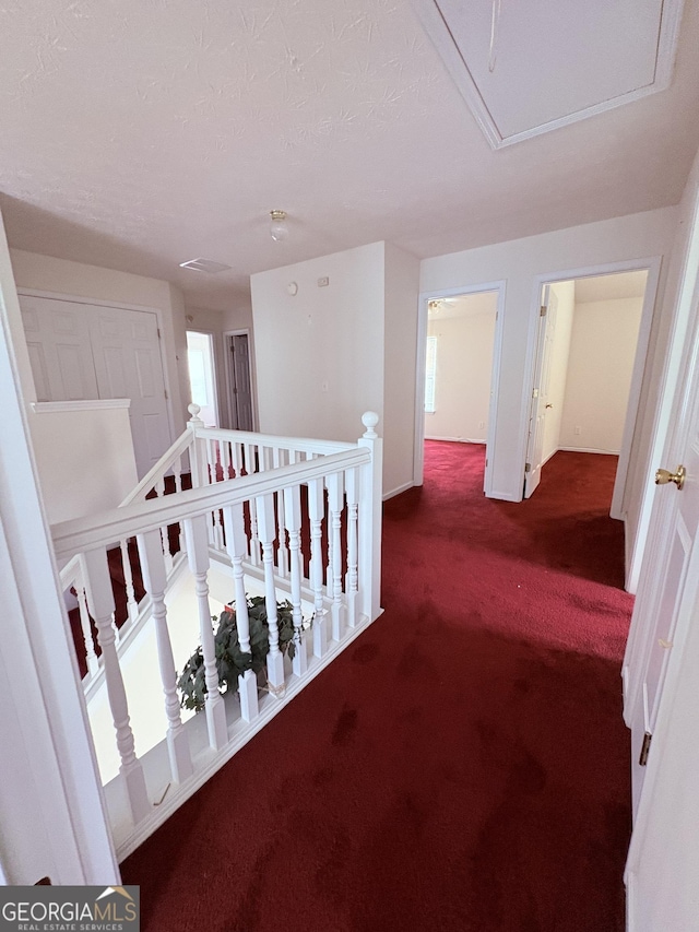 hall with carpet flooring, a textured ceiling, attic access, baseboards, and an upstairs landing
