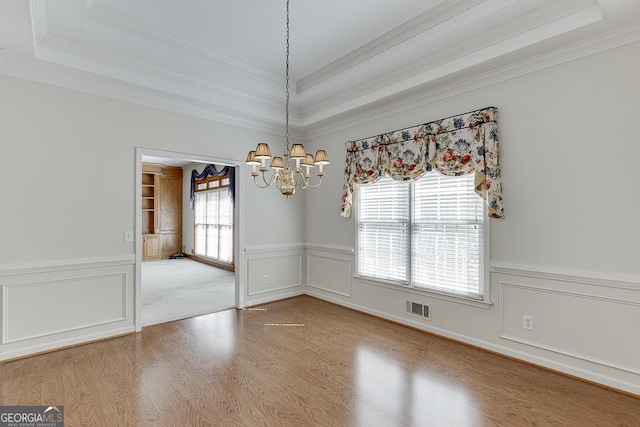 unfurnished dining area with wood finished floors, a chandelier, crown molding, a raised ceiling, and a wainscoted wall