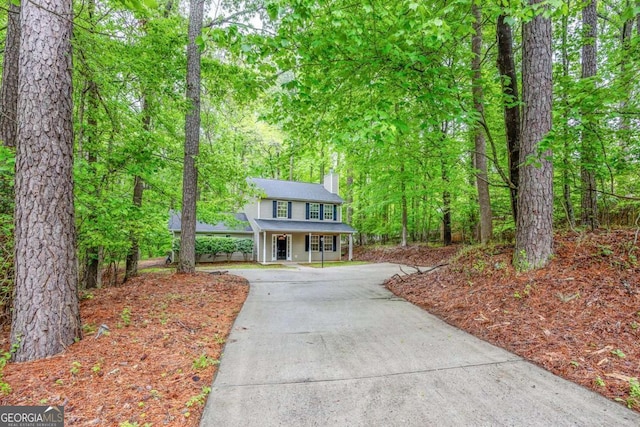 view of front facade with covered porch, a view of trees, a chimney, and concrete driveway