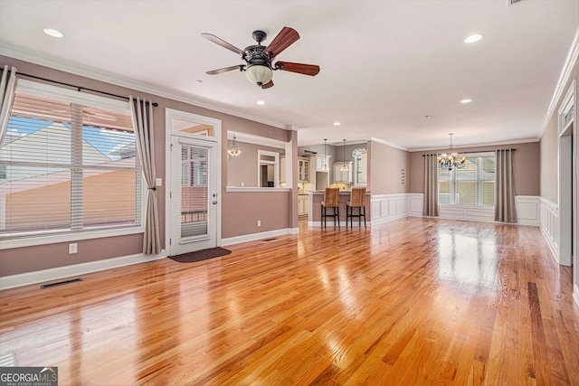 unfurnished living room with ornamental molding, a chandelier, a ceiling fan, light wood-style flooring, and wainscoting
