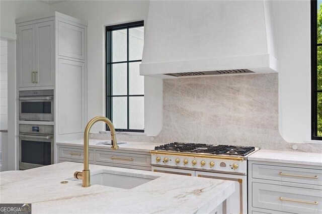 kitchen with a sink, tasteful backsplash, custom exhaust hood, range with two ovens, and light stone countertops