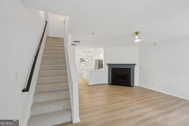unfurnished living room with a fireplace with flush hearth, light wood-type flooring, baseboards, a ceiling fan, and stairs