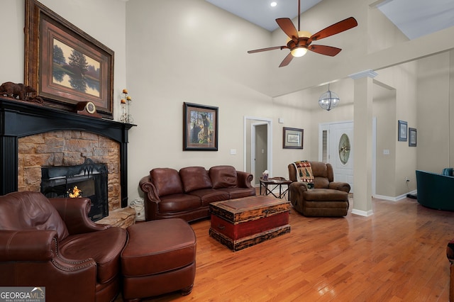 living area with ceiling fan, a stone fireplace, light wood finished floors, baseboards, and a towering ceiling