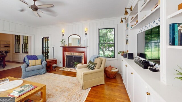 living area featuring light wood-style flooring, ceiling fan, crown molding, and a brick fireplace