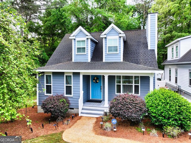 cape cod-style house featuring roof with shingles, a porch, and a chimney