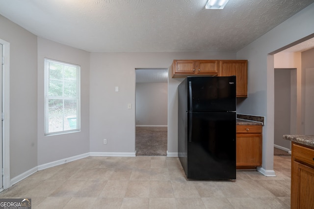 kitchen featuring brown cabinets, a textured ceiling, freestanding refrigerator, dark stone counters, and baseboards