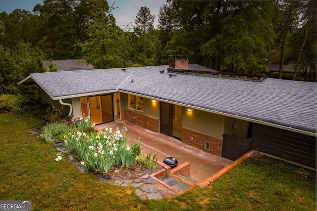 rear view of house with a chimney, roof with shingles, a patio area, and a lawn