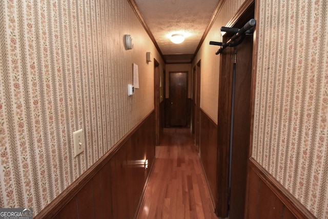 hall with a textured ceiling, wallpapered walls, wainscoting, and wood finished floors