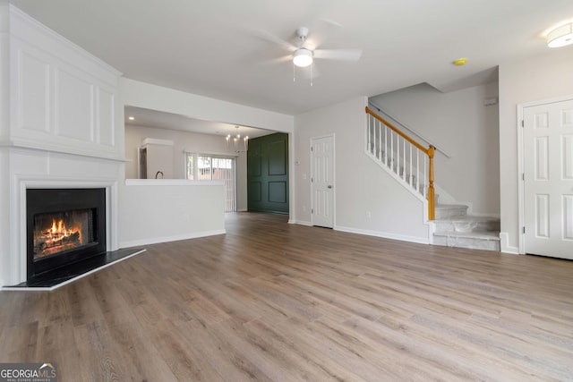 unfurnished living room featuring ceiling fan with notable chandelier, wood finished floors, stairway, baseboards, and a glass covered fireplace