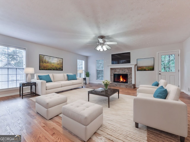 living room with a textured ceiling, ceiling fan, baseboards, light wood finished floors, and a stone fireplace