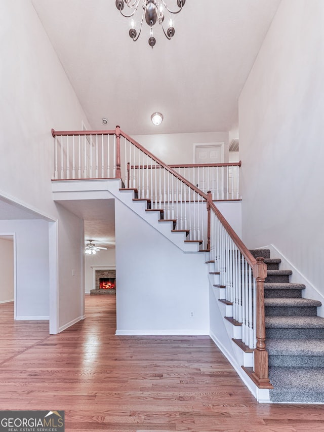 staircase featuring wood finished floors, ceiling fan with notable chandelier, baseboards, a lit fireplace, and a towering ceiling