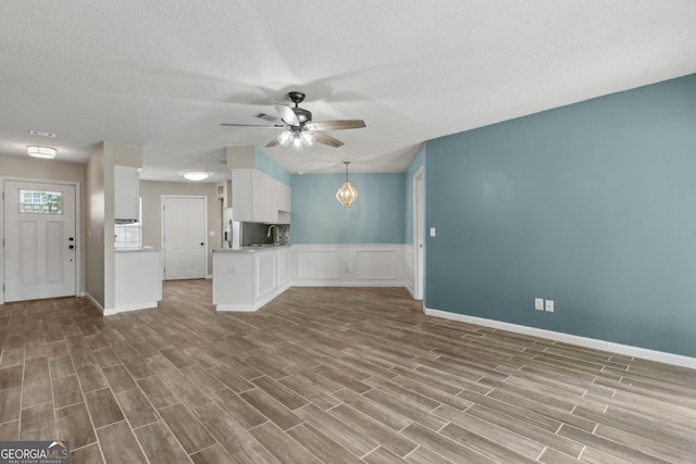 unfurnished living room featuring a textured ceiling, ceiling fan, baseboards, wood finished floors, and wainscoting