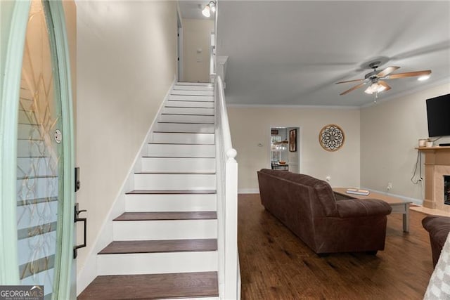staircase with baseboards, ornamental molding, a fireplace, wood finished floors, and a ceiling fan