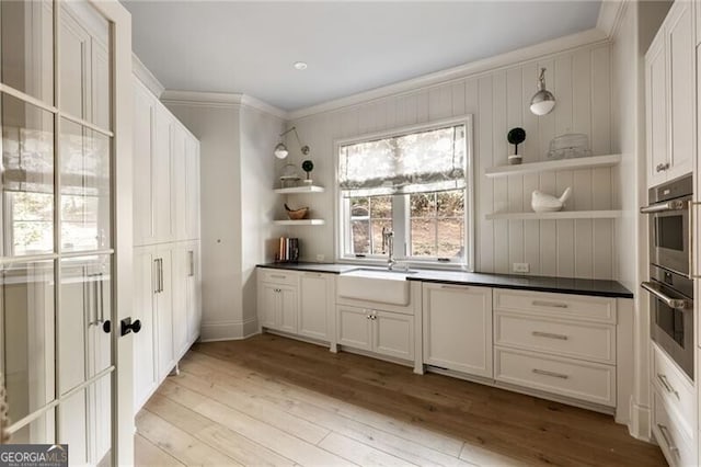 kitchen with crown molding, open shelves, light wood-style floors, stainless steel double oven, and a sink