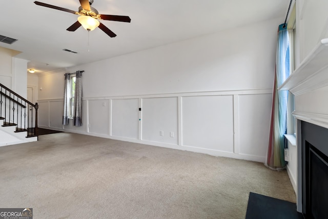 unfurnished living room with a decorative wall, a ceiling fan, stairs, carpet, and wainscoting