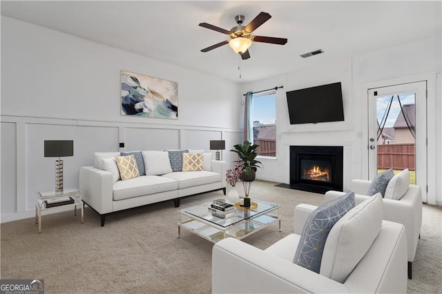 living room with carpet flooring, a ceiling fan, a decorative wall, a fireplace with flush hearth, and wainscoting