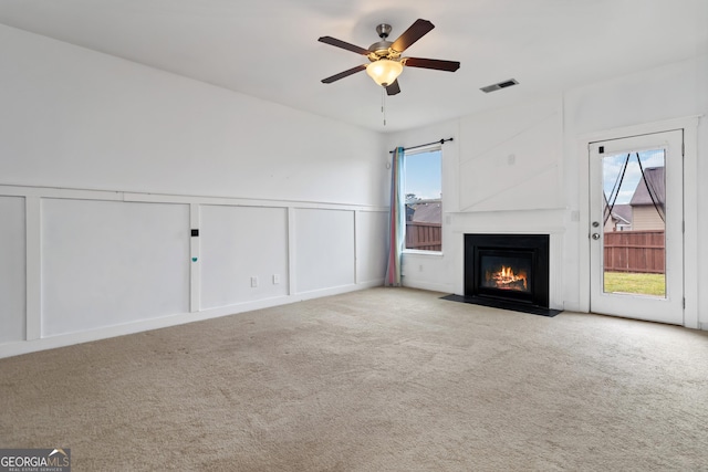 unfurnished living room featuring carpet, ceiling fan, a decorative wall, a fireplace with flush hearth, and wainscoting