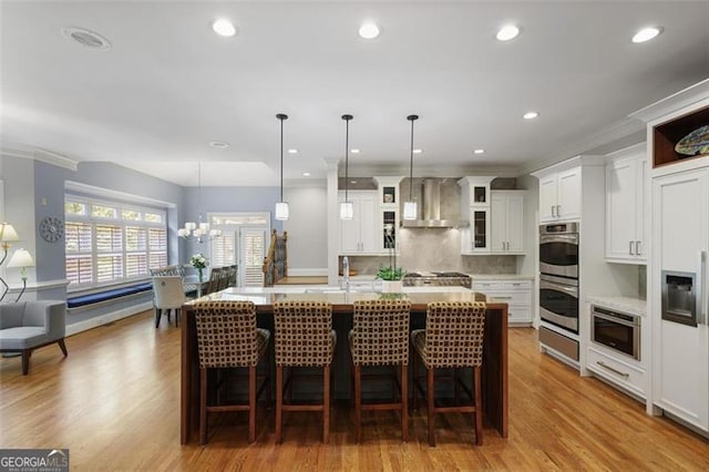 kitchen featuring decorative backsplash, a kitchen island with sink, double oven, wall chimney range hood, and white cabinets