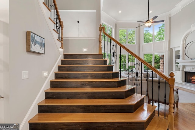staircase with ceiling fan, crown molding, a premium fireplace, wood finished floors, and a high ceiling
