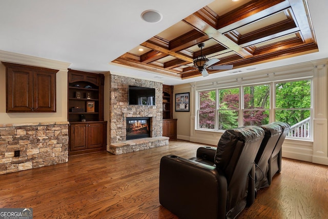 living area with ornamental molding, a ceiling fan, a stone fireplace, coffered ceiling, and wood finished floors