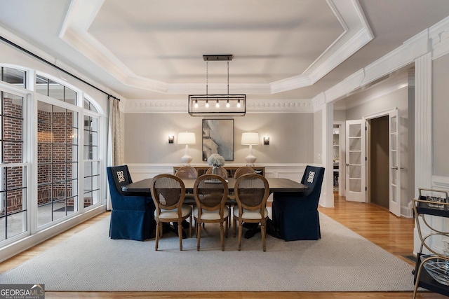 dining room featuring a tray ceiling, crown molding, light wood-style flooring, a decorative wall, and wainscoting