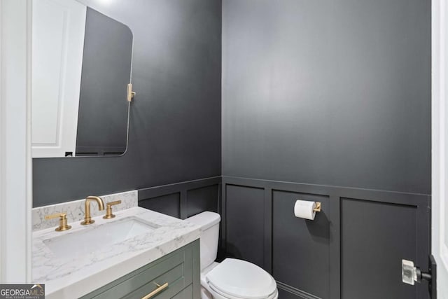 half bathroom with a decorative wall, toilet, a wainscoted wall, and vanity