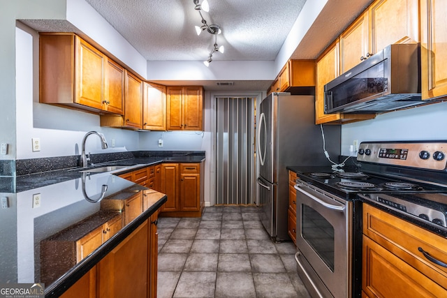kitchen with a textured ceiling, brown cabinets, a sink, dark stone countertops, and appliances with stainless steel finishes