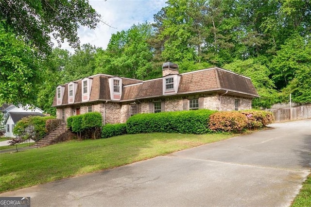 view of front facade with a front lawn, a shingled roof, mansard roof, a chimney, and fence