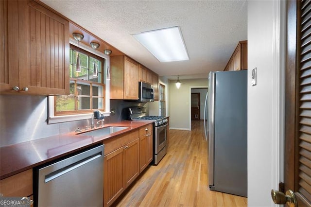 kitchen with a textured ceiling, a sink, appliances with stainless steel finishes, light wood-type flooring, and brown cabinetry