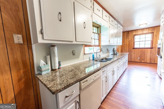 kitchen featuring white dishwasher, a sink, white cabinetry, light wood-type flooring, and wooden walls