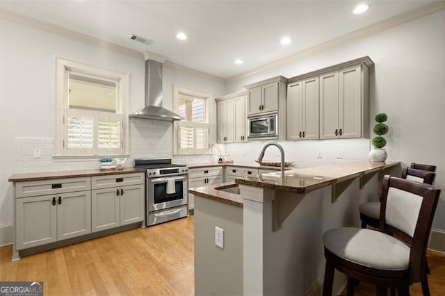 kitchen with crown molding, appliances with stainless steel finishes, a kitchen breakfast bar, wall chimney exhaust hood, and a peninsula